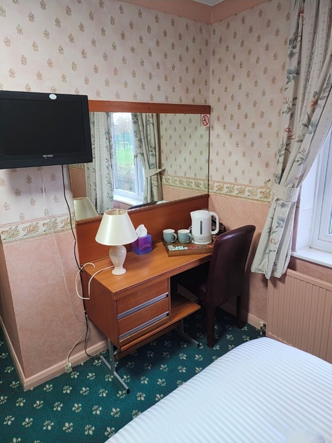 Une chambre d’hôtel équipée d’un lit, d’un bureau avec une lampe, d’une machine à café, d’un miroir et d’un téléviseur fixé au mur.