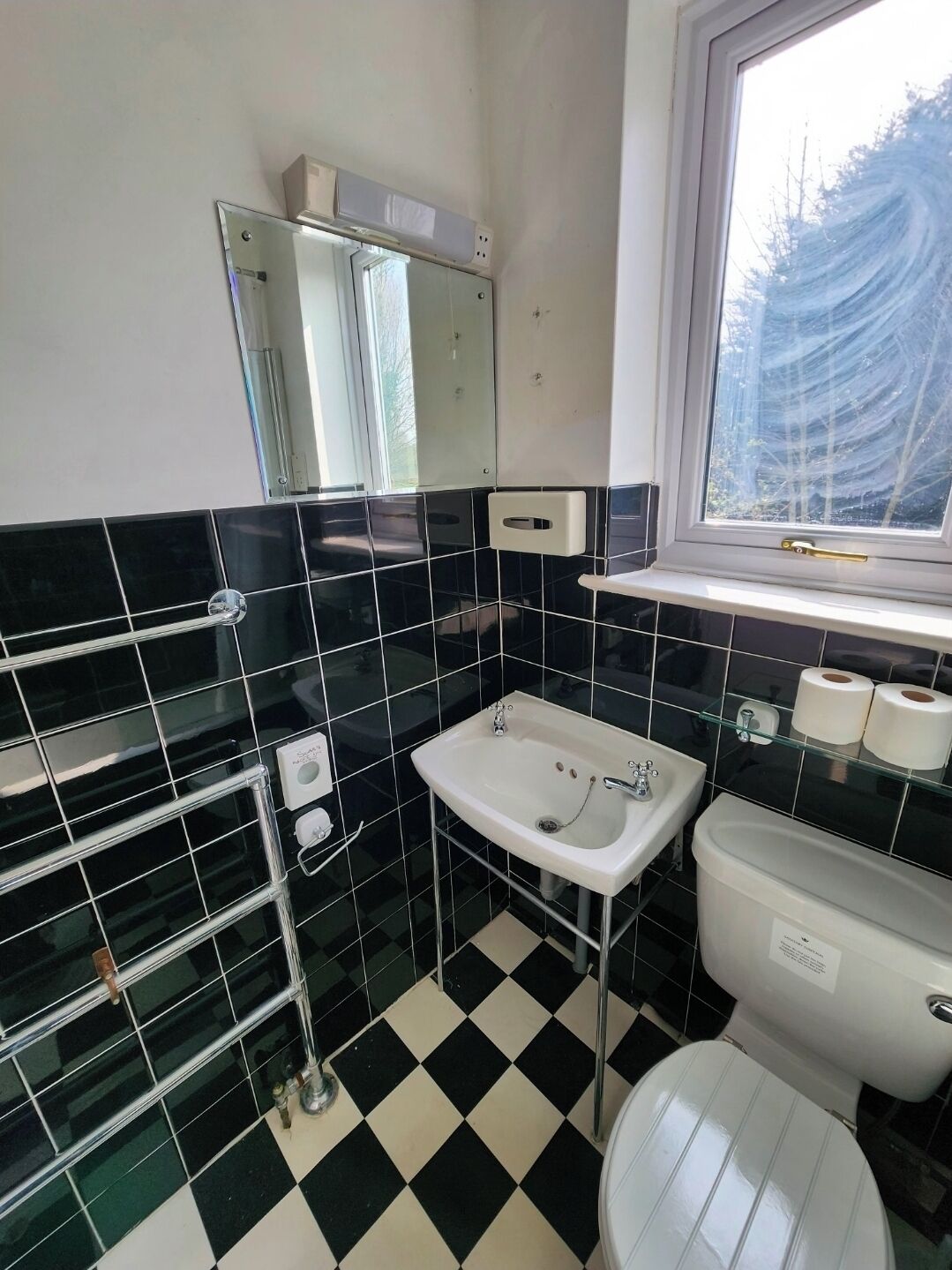 Ein Badezimmer mit Schachbrettboden, einer weißen Toilette, einem Waschbecken und einem Fenster mit Blick auf Bäume.