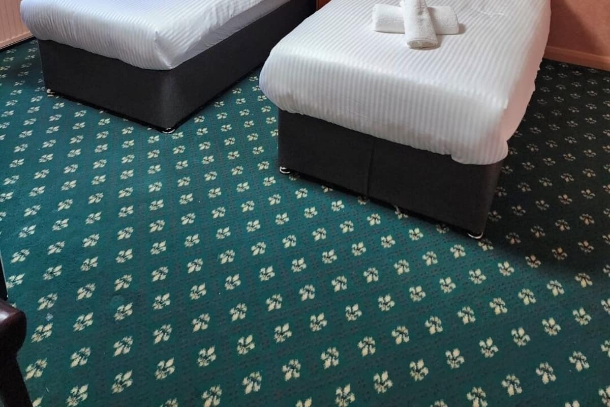 Una habitación de hotel con dos camas, cabecera de madera, alfombra estampada y cortina con motivos florales.