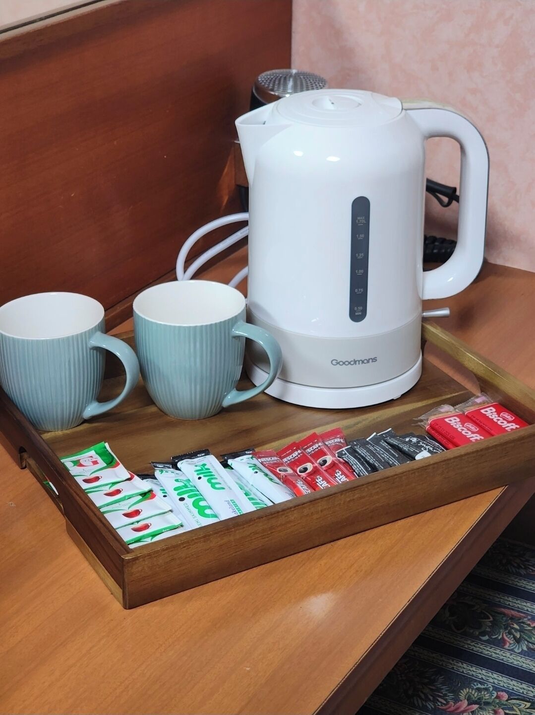 A white electric kettle, two light blue mugs, and a tray with tea and biscuit packets.