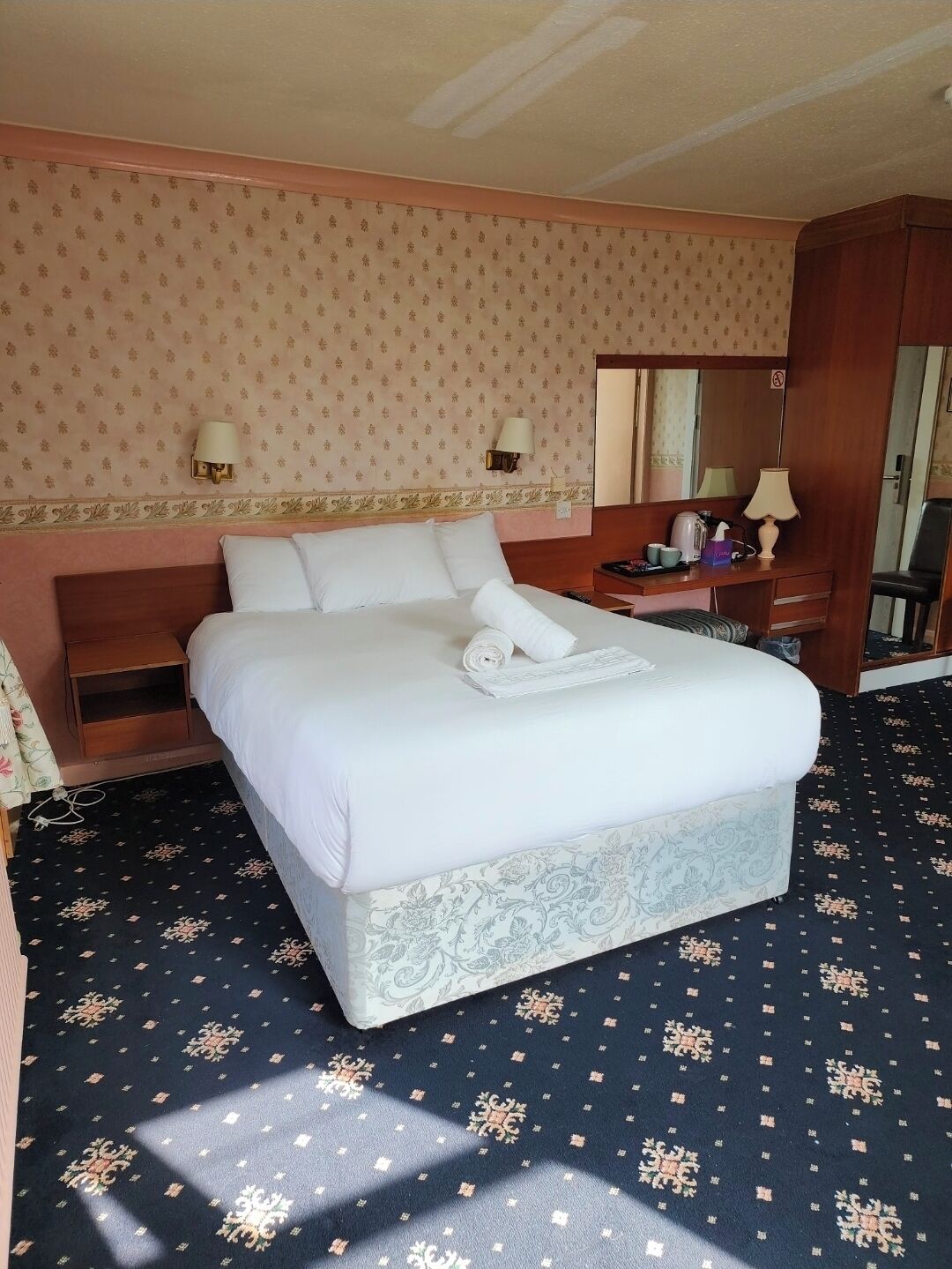 Una camera d'albergo con un letto grande, un comodino in legno, una scrivania con una lampada e uno specchio.