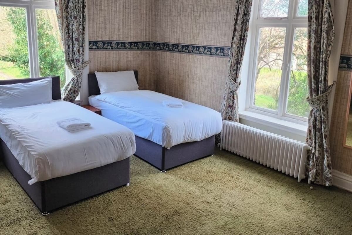 Habitación de hotel con dos camas, un radiador y ventanas grandes con cortinas.