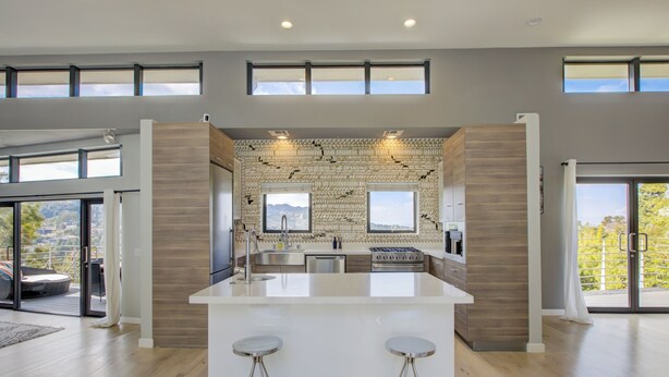 A modern kitchen with a central island, two bar stools, and a view of the outdoors through large windows. - The Hollywood Hills Executive - Incredible Sunsets- Next to Studios! (Los Angeles)