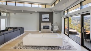 A modern living room with a fireplace, a large sofa, a coffee table, and a balcony with outdoor seating. - The Hollywood Hills Executive - Incredible Sunsets- Next to Studios! (Los Angeles)