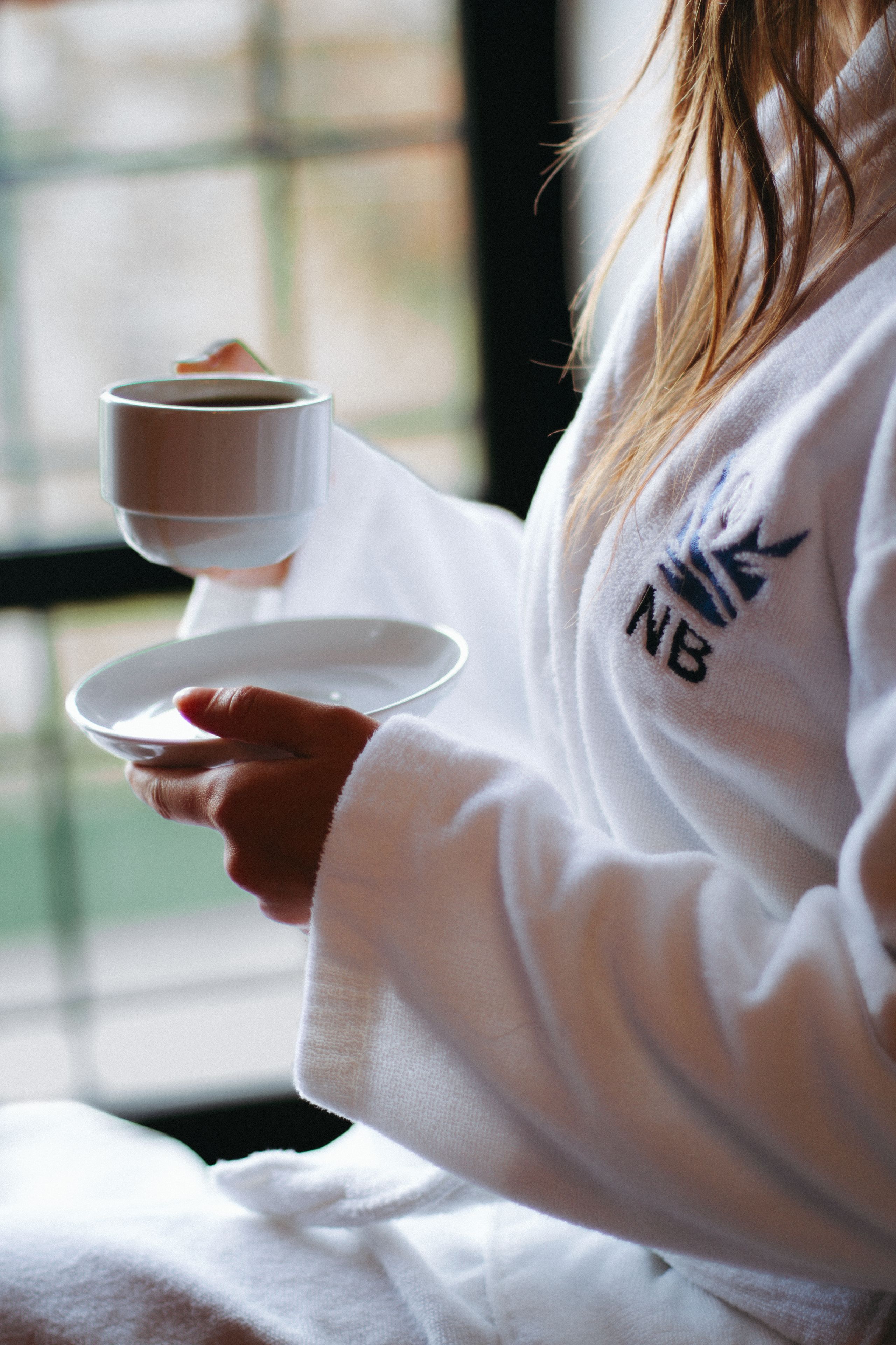 A person holding a cup and saucer, wearing a white robe with a logo, sitting by a window.