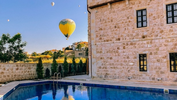 A stone building with a swimming pool, hot air balloons in the background, and a clear sky. - Noble Blue Cappadocia  (Urgup)