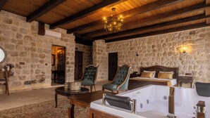 A stone-walled room with a large bathtub, two upholstered chairs, a wooden table, and a chandelier. - Noble Blue Cappadocia  (Urgup)