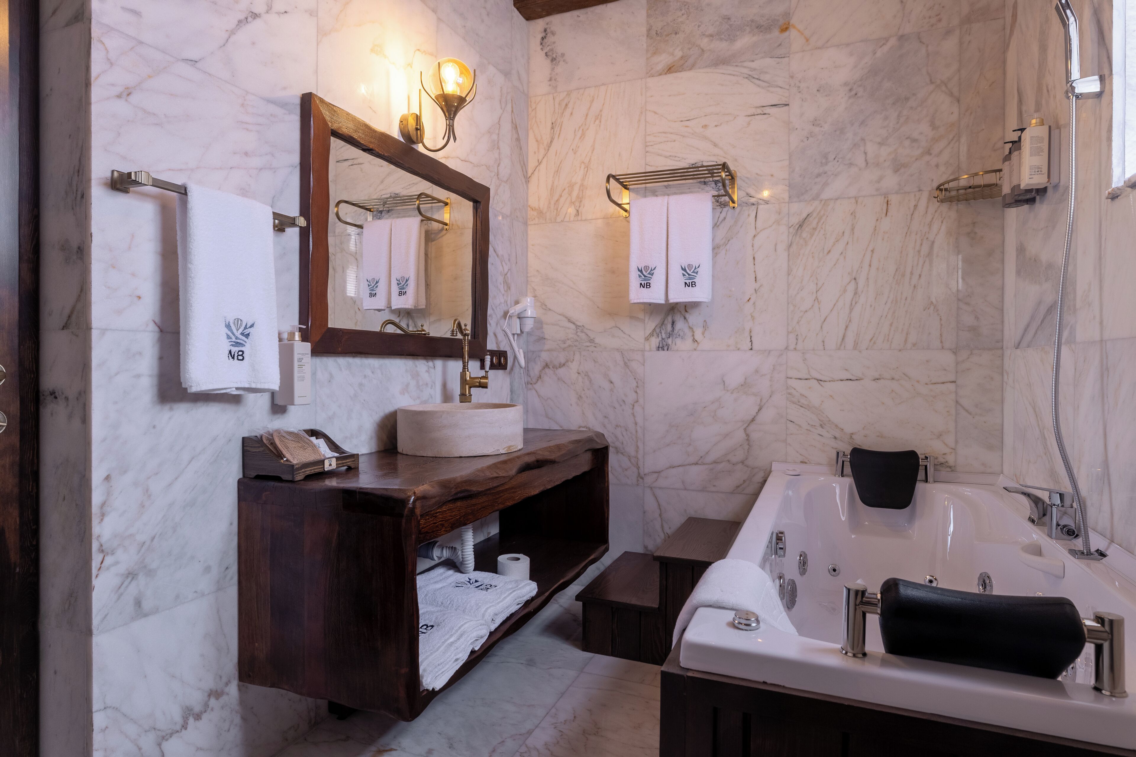 A bathroom with a marble wall, a wooden vanity, a large bathtub, and a mirror with wall-mounted lights.