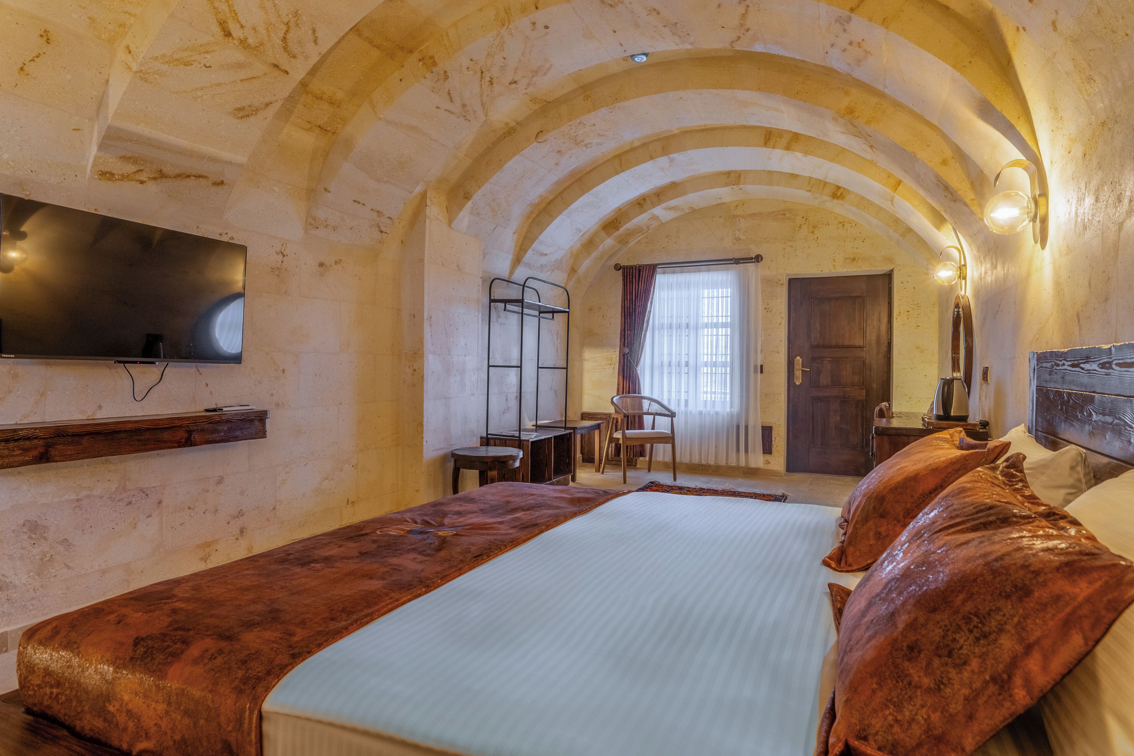 A room with a bed, a flat-screen TV mounted on the wall, a wooden bench, and a door leading to another room.