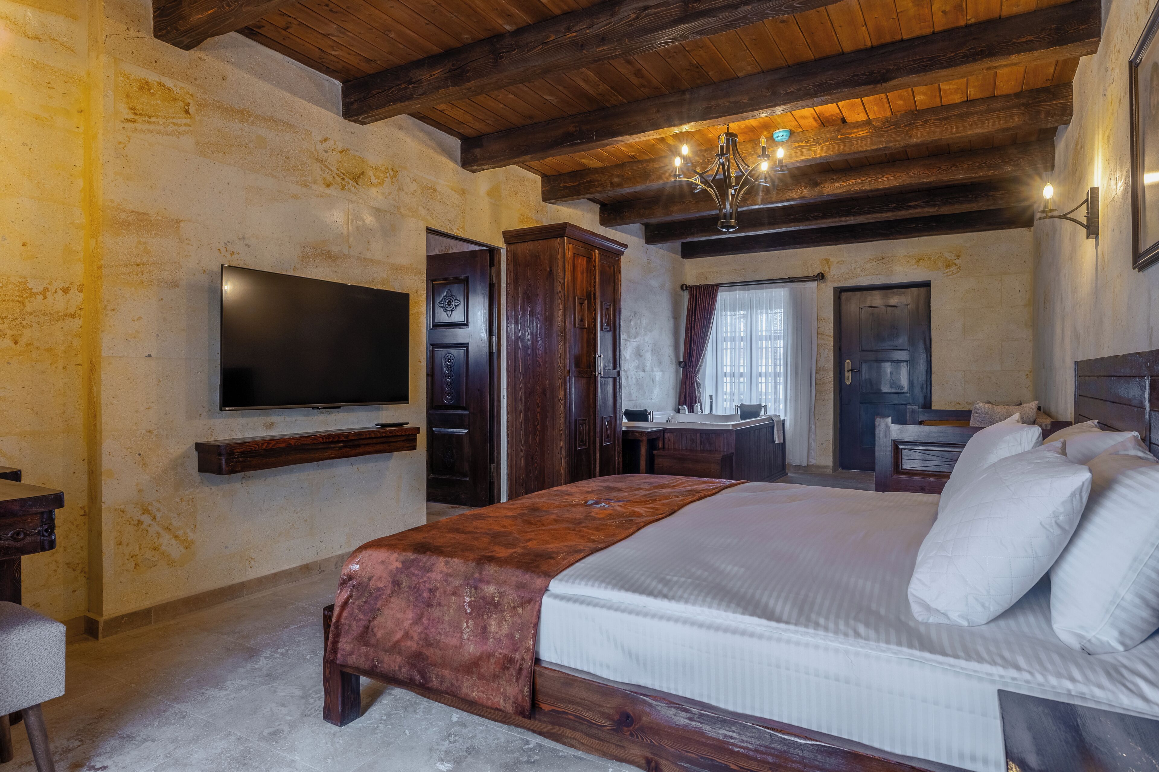 A bedroom with a large bed, a television mounted on the wall, a wooden ceiling, and a chandelier.