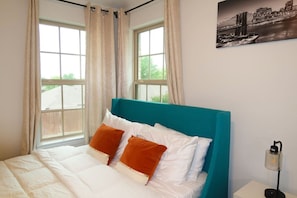 A bedroom with a bed, two windows with curtains, a teal headboard, and a cityscape painting on the wall.