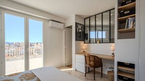Ein Schlafzimmer mit einem Bett, einem Schreibtisch, einem Stuhl, einem Bücherregal und einem großen Fenster mit Blick auf die Stadt.