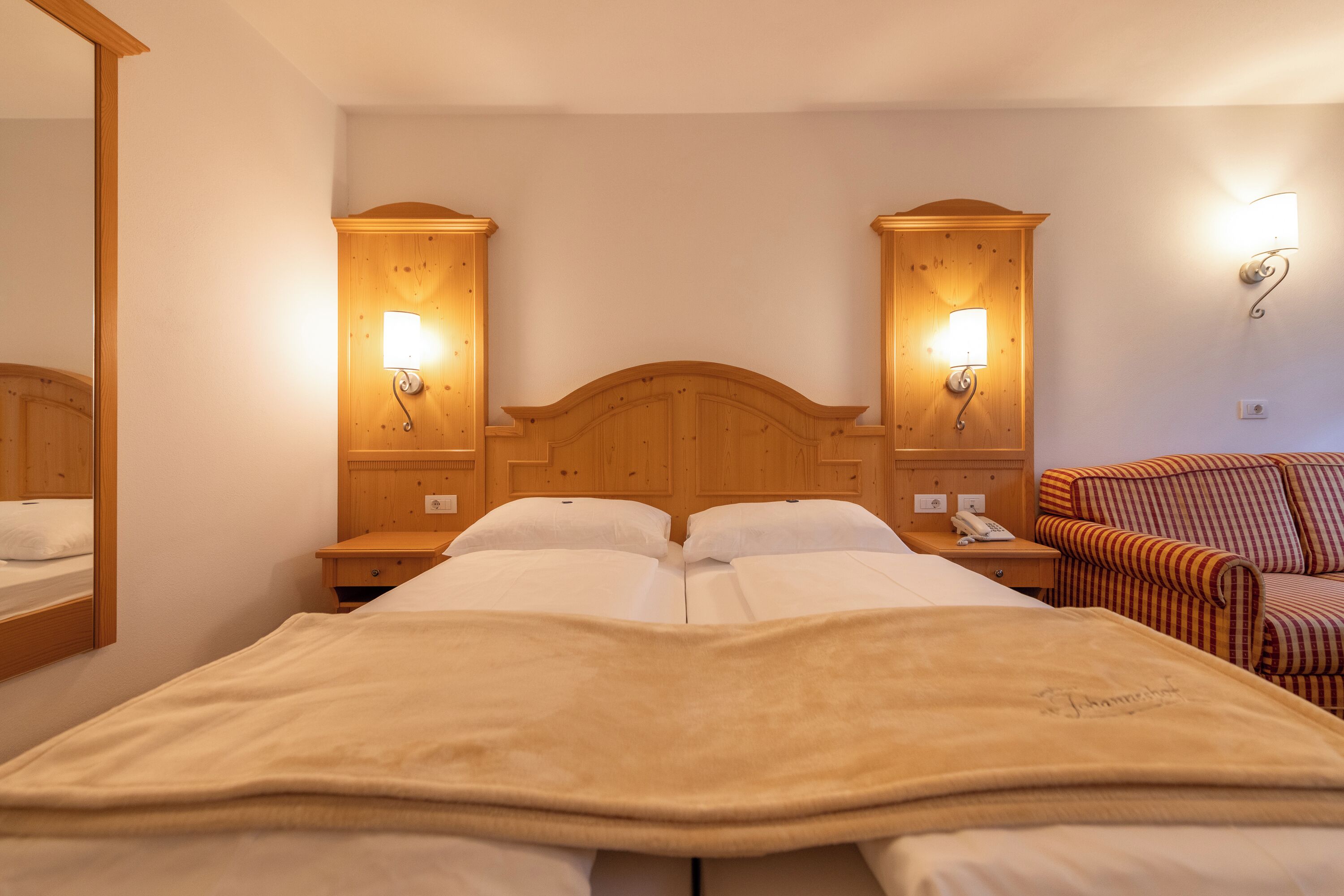 Un lit double avec une couverture beige, une tête de lit en bois, des lampes de chevet, une table de chevet avec un téléphone et un fauteuil à rayures.