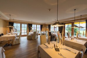 A restaurant with multiple tables set for dining, large windows, and hanging pendant lights. - Berghotel Johanneshof (Rasun Anterselva)