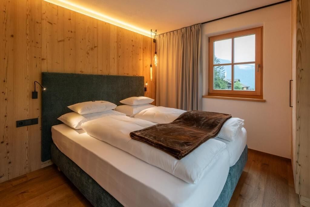 A bedroom with a wooden wall, a bed with white linens, a brown throw blanket, and a window with curtains.