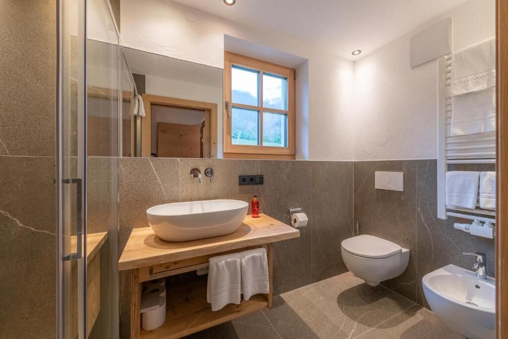 A bathroom with a large vessel sink, a toilet, a bidet, a glass shower enclosure, a wooden vanity, and a window with a view.