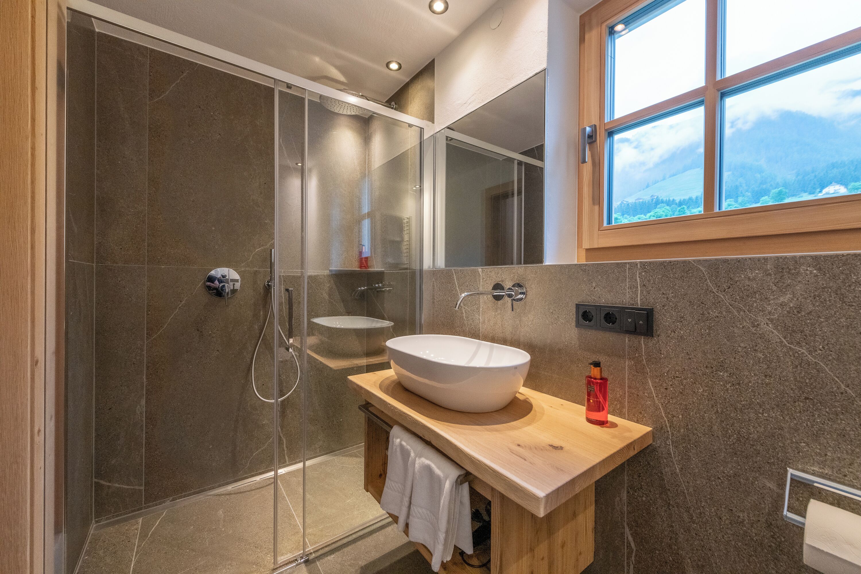 Un baño moderno con cabina de ducha de vidrio, tocador de madera con un lavamanos grande tipo recipiente y ventana con vistas a la montaña.