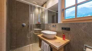 Un baño moderno con cabina de ducha de vidrio, tocador de madera con un lavamanos grande tipo recipiente y ventana con vistas a la montaña.
