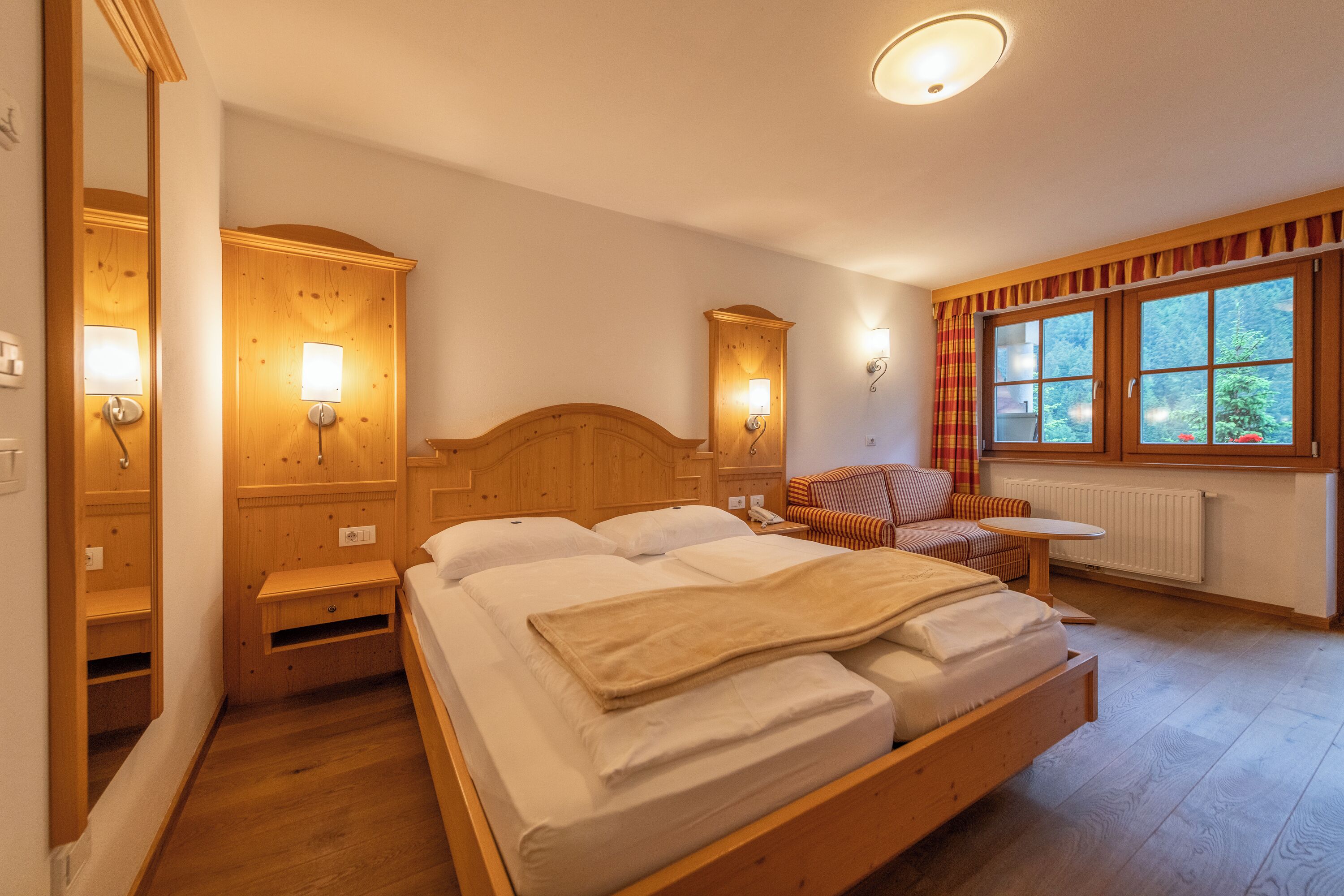 A bedroom with a wooden bed, bedside tables, a chair, a small table, and a window with curtains.