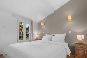 Ein Schlafzimmer mit einem großen Bett, zwei Nachttischlampen, einem Fenster mit Blick ins Grüne und einer Tür.