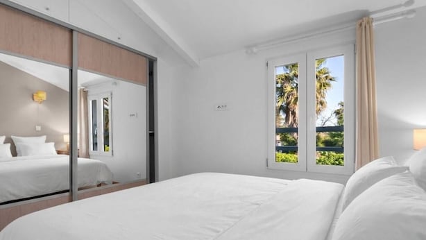 Hotellihuone, jossa on suuri sänky, peili, ikkuna, josta avautuu näkymä palmuille, ja ovi, joka johtaa parvekkeelle.