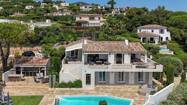 Una casa de varios pisos con piscina, rodeada de exuberante vegetación y otras viviendas.