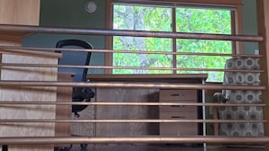 A cabin interior with a wooden ceiling, a desk with a chair, a window with a view of trees, and a patterned cushion on a bench. - Heron's Nest - Elegant King Master with custom woodwork, lofts and stained glass (Eugene)