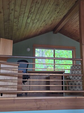 A cabin interior with a wooden ceiling, a desk with a chair, a window with a view of trees, and a patterned cushion on a bench. - Heron's Nest - Elegant King Master with custom woodwork, lofts and stained glass (Eugene)