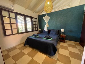 A bedroom with a bed, two bedside tables, a window with blinds, a hanging light fixture, and a patterned wall.