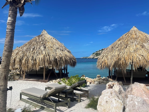 A beachside resort with thatched-roof structures, lounge chairs, and a clear view of the sea.