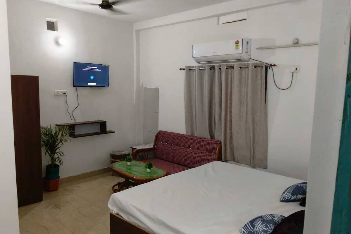 Ein Schlafzimmer mit einem Bett, einem Fernseher, einem Deckenventilator, einer Klimaanlage, einem Fenster mit Vorhängen und einem kleinen Tisch mit einer Pflanze.