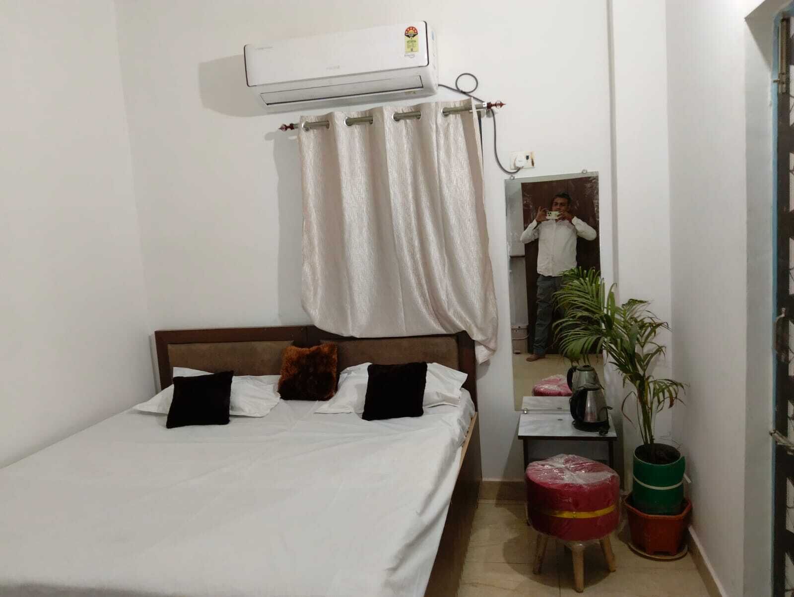 A single bed with white bedding, a wooden headboard, a small bedside table with a kettle, and a potted plant.