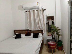 Una cama individual con ropa blanca, un cabecero de madera, una mesita de noche con una tetera y una planta en maceta.
