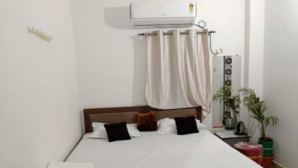 Een bed met witte beddengoed, een houten hoofdbord en een wit gordijn. Er staat een klein tafeltje met een waterkoker en een plantje.