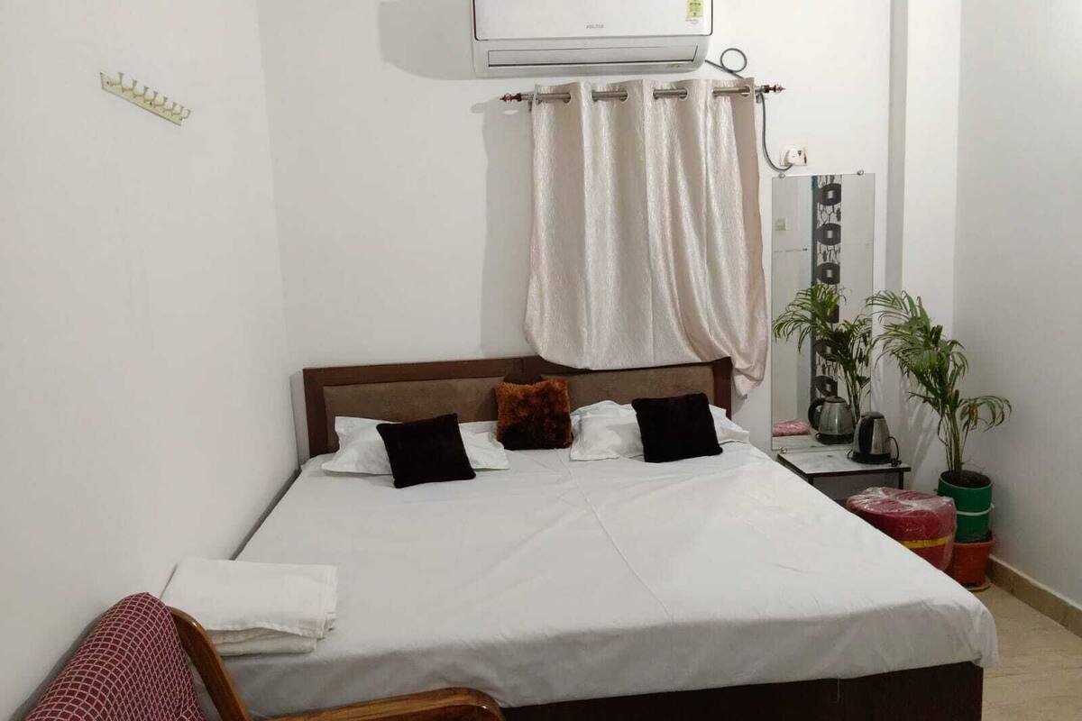 Ein Bett mit weißer Bettwäsche, einem Holzkopfschild und einem weißen Vorhang. Es steht ein kleiner Tisch mit einem Wasserkocher und einer Pflanze daneben.