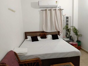 Ein Bett mit weißer Bettwäsche, einem Holzkopfschild und einem weißen Vorhang. Es steht ein kleiner Tisch mit einem Wasserkocher und einer Pflanze daneben.