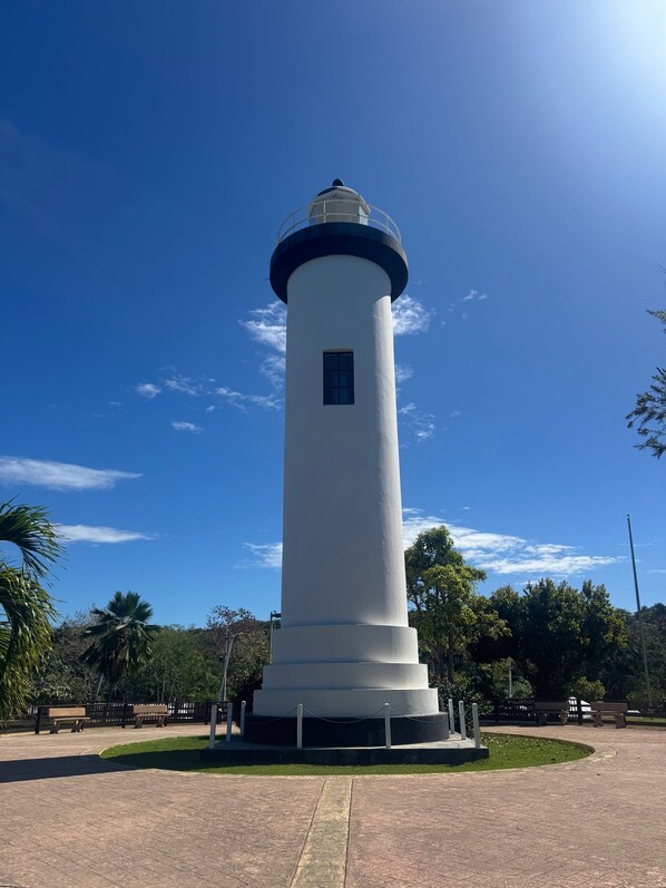 Un faro alto y blanco con la parte superior azul, rodeado de un área pavimentada con bancos y vegetación.