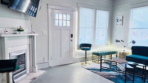 En stue med pejs, tv monteret på væggen, sofa, bænk og et vindue med skodder.