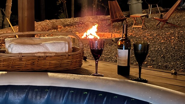 An outdoor hot tub with a fire, a wicker basket with bread, and a bottle of wine.