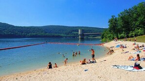 A beach with people swimming and sunbathing, a bridge in the background, and a forested hillside.