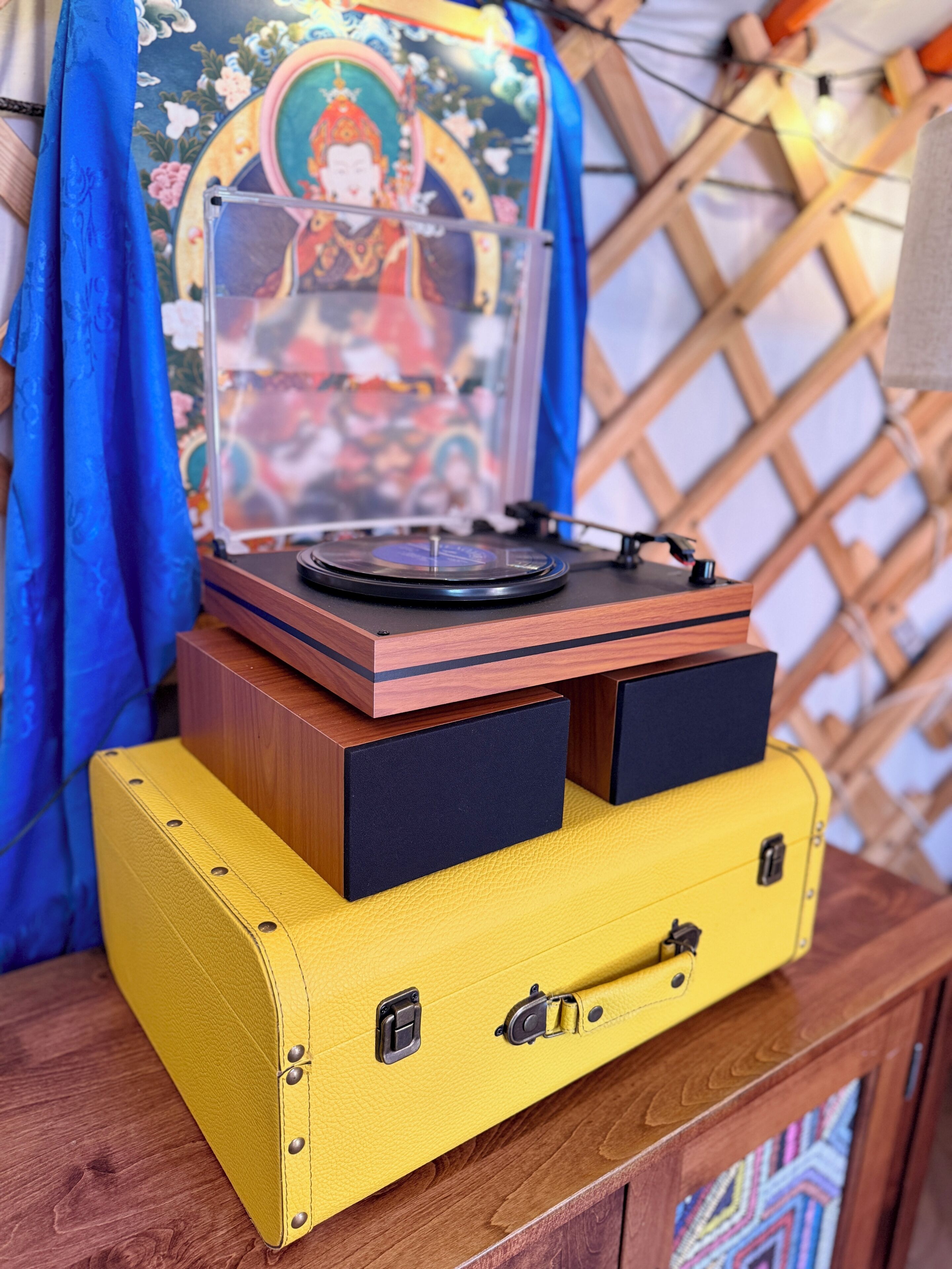 Uma mesa de madeira com um toca-discos, uma mala amarela e uma tapeçaria colorida ao fundo.