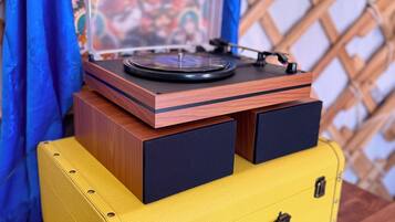 Uma mesa de madeira com um toca-discos, uma mala amarela e uma tapeçaria colorida ao fundo.