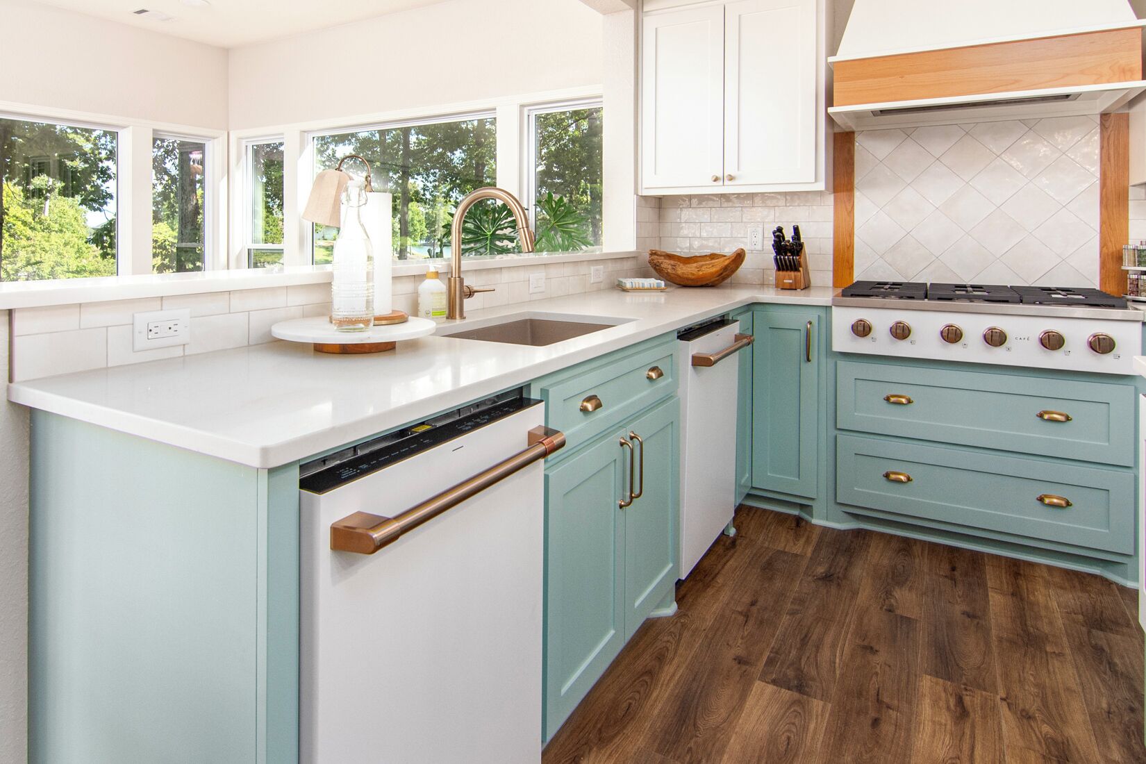 A kitchen with white and teal cabinetry, a large island with a sink, a stove, and a view of greenery outside.