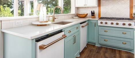 A kitchen with white and teal cabinetry, a large island with a sink, a stove, and a view of greenery outside.