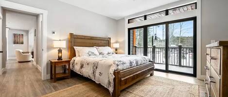 Un dormitorio con una cama grande, dos mesitas de noche con lámparas, un cabecero de madera, un ventanal con vista a árboles y un balcón con barandilla.