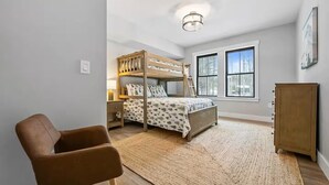 Ein Schlafzimmer mit Etagenbett, Schreibtisch, Stuhl, Kleiderschrank und großen Fenstern.