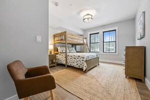 Ein Schlafzimmer mit Etagenbett, Schreibtisch, Stuhl, Kleiderschrank und großen Fenstern.
