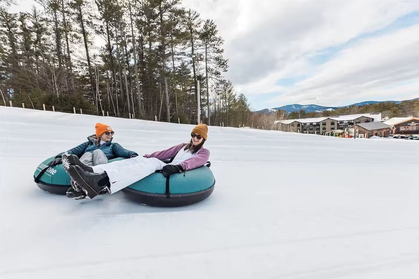 Two people on snow tubes on a snowy slope with buildings in the background.