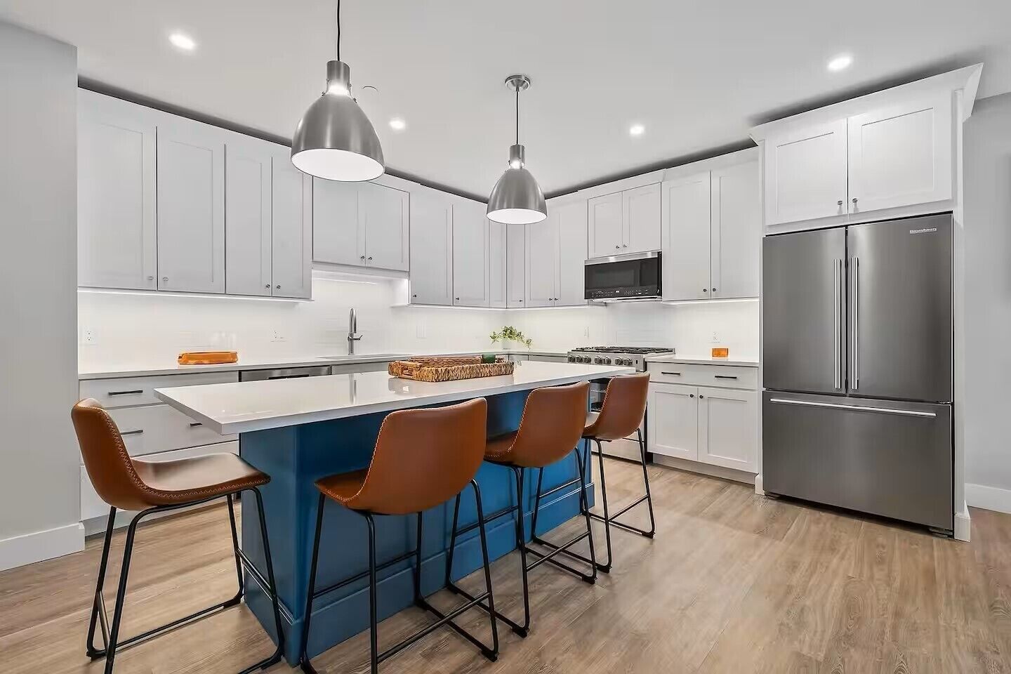 A modern kitchen with white cabinets, a stainless steel refrigerator, a central island with bar stools, and pendant lighting.