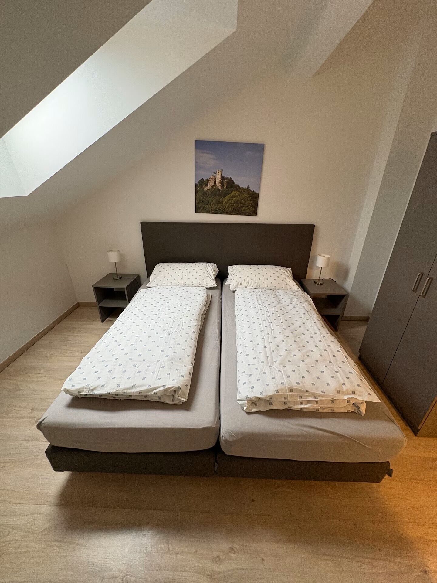 Een kamer met twee bedden, een nachtkastje, een lamp, een kledingkast en een schilderij aan de muur.